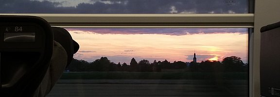 Durch das Zugfenster eines schnell fahrenden Zuges sind man in der Ferne die Silhouette eines Kirchturms. 